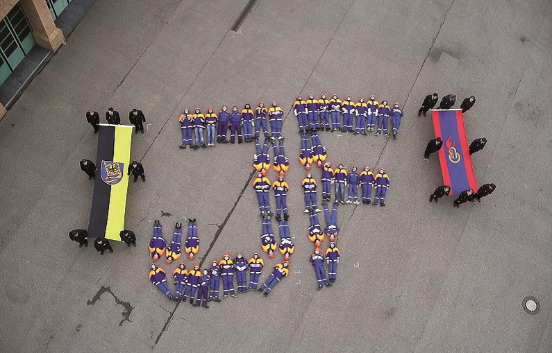 Foto aus der Vogelperspektive: Mitglieder der Jugendfeuerwehr liegen in Formation der Initialen JF auf dem Boden des Feuerwehrhofes